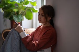 Sad depressed teen adolescent girl looking side hold phone sit on floor at home. Upset teenager with cellphone waiting for call feel bad anxious need help. Cyberbullying, difficult relations at school