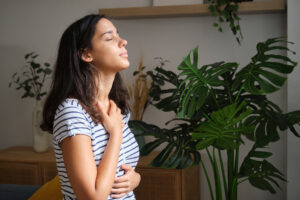 Young woman is taking a deep breath while standing in her living room, surrounded by plants
