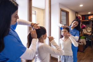 A Latina nurse in uniform brushing her daughter's hair while the girl brushes her teeth in front of the mirror. A heartfelt morning moment showing love, care, and responsibility before starting a day of work in healthcare.