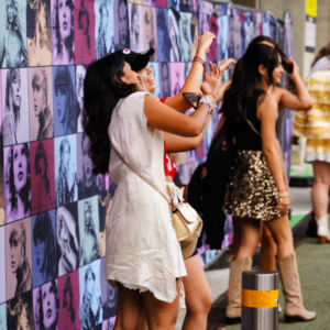 Four teen girls pose in front of a multicolor photo backdrop of Taylor Swift at Taylor Swift's Eras tour. Swifties are one of the largest fandoms in the world.