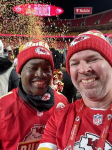 Matt Arnet (right) and a Chiefs Rumble team member pose on GEHA Field at Arrowhead Stadium in front of yellow confetti spraying in the air behind them