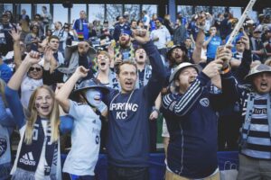 A group of Sporting Kansas City soccer fans all cheering in the stands as they all wear shared of navy to light blue